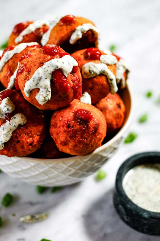 Angled shot of buffalo chicken meatballs in a bowl with ranch and hot sauce drizzled on top. Extra ranch and chopped green onion on the side. 