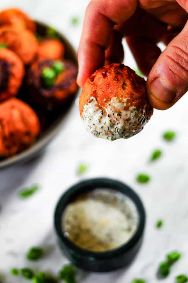 Buffalo chicken meatballs being dipped into ranch sauce. Bowl of meatballs in the background with chopped green onion garnish. 