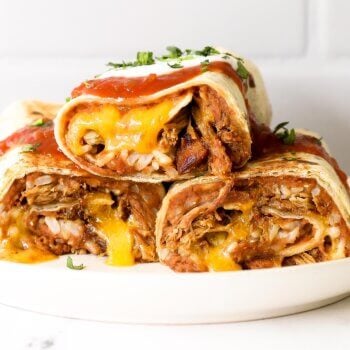 Straight on shot of 3 baked carnitas burritos on a plate with salsa and sour cream on top