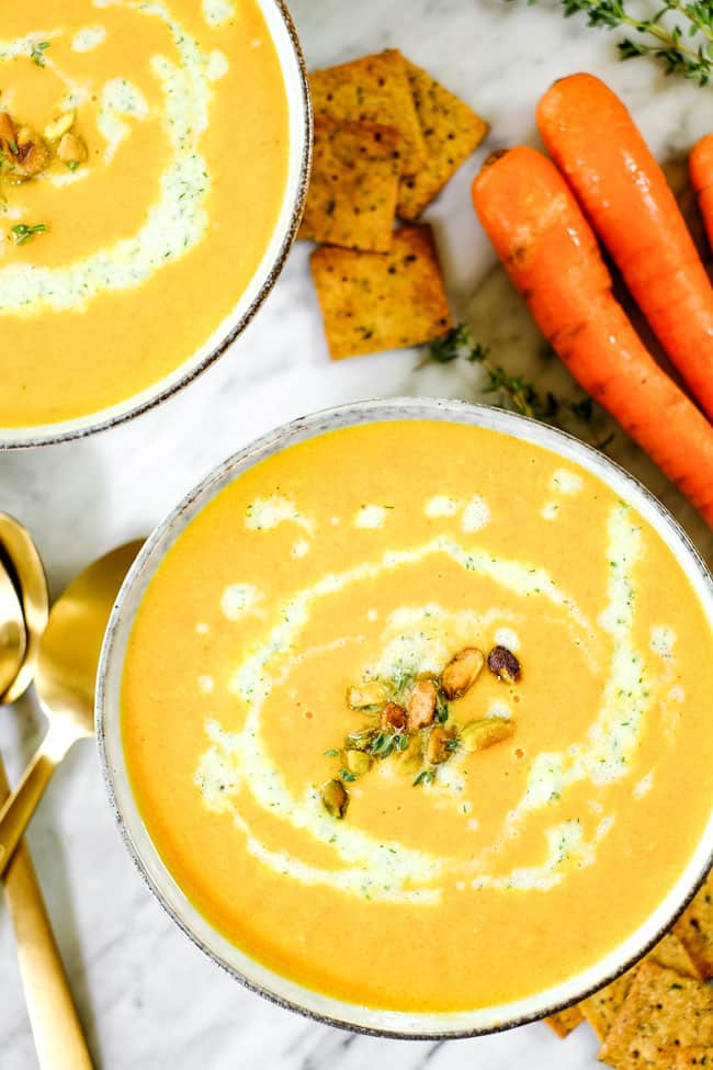 Carrot ginger soup in two bowls with tzatziki sauce, pistachios and fresh thyme. On a table with extra sauce, thyme, crackers and carrots. 