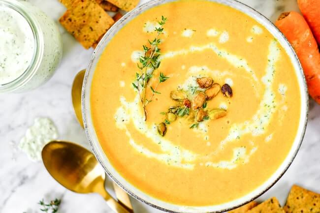 Carrot ginger soup with tzatziki sauce, pistachios and fresh thyme. On a table with extra sauce, crackers and carrots. 