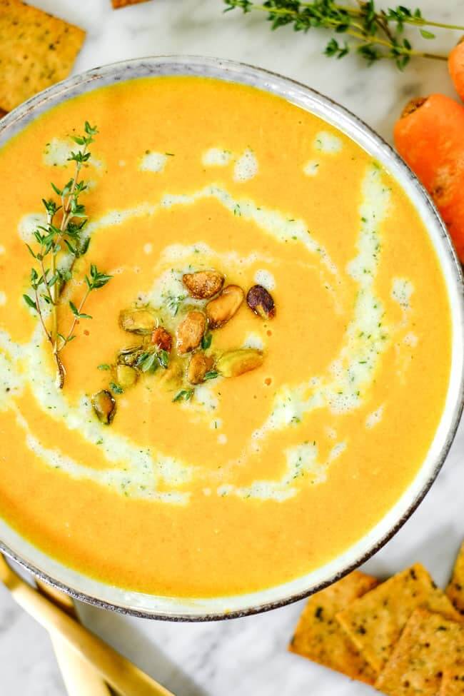 Close up shot of carrot ginger soup with tzatziki sauce, pistachios and fresh thyme. On a table with extra sauce, thyme, crackers and carrots. 