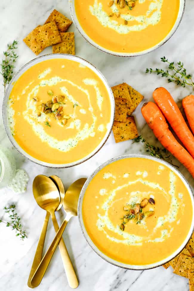 Carrot ginger soup in three bowls with tzatziki sauce, pistachios and fresh thyme. On a table with extra sauce, thyme, crackers and carrots. 