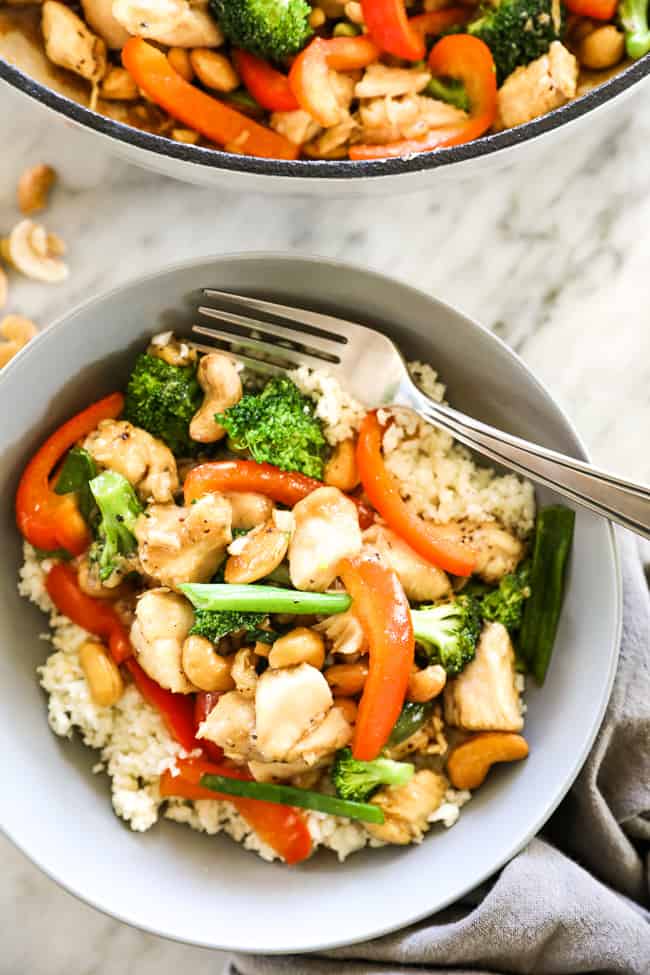Overhead close up image of cashew chicken served in a bowl over cauliflower rice with a fork. Chicken, cashews, broccoli, red bell pepper and an asian sauce. 