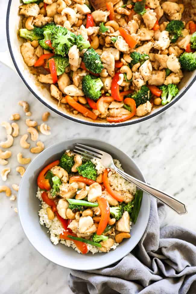 Overhead image of cashew chicken in a skillet and in a bowl served over cauliflower rice with a fork. Chicken, cashews, broccoli, red bell pepper and an asian sauce. 