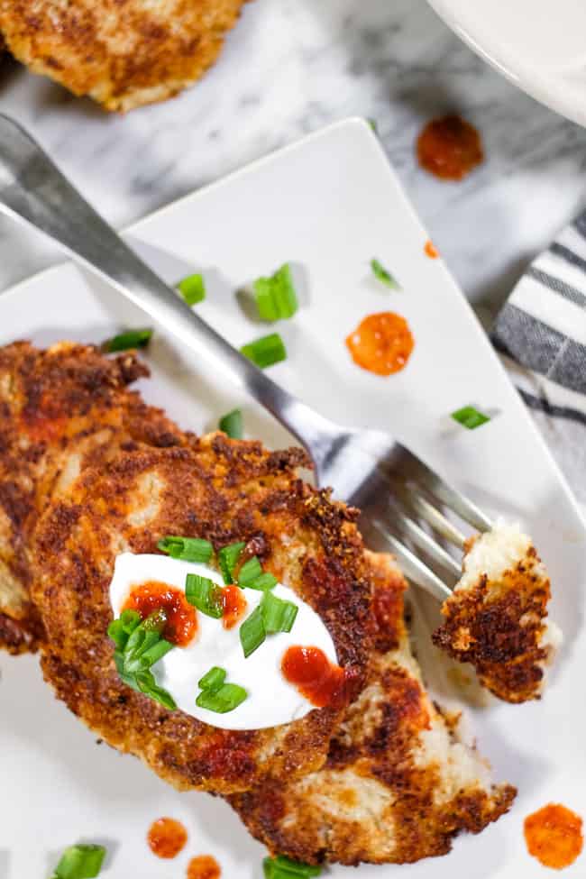 Close up of cauliflower fritters on a plate with a bit taken out on a fork. Topped with hot sauce, coconut yogurt and chopped green onion. 