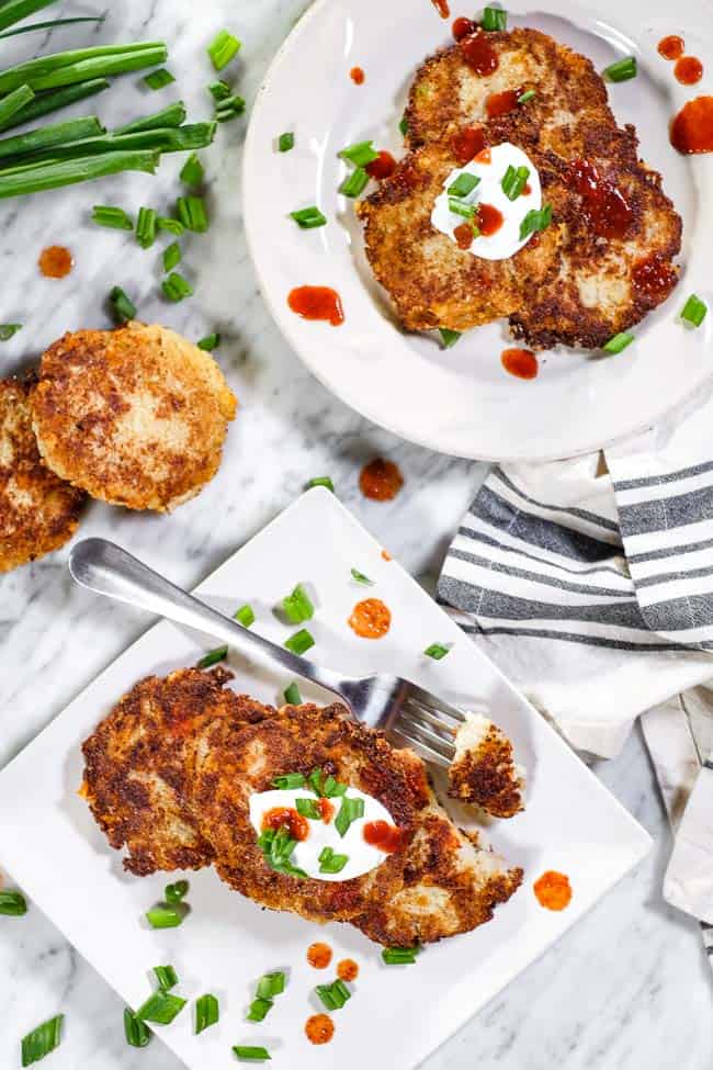 Cauliflower fritters laid on on two plates, topped with hot sauce, coconut yogurt and chopped green onion. One pate has a bite taken out on a fork. 