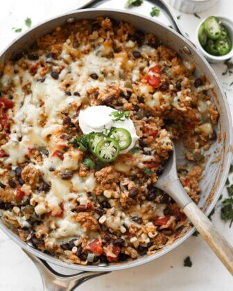 Skillet filled with cheesy rice, black beans, corn, tomatoes, and ground meat, topped with sour cream and sliced jalapeños, served with a wooden spoon.