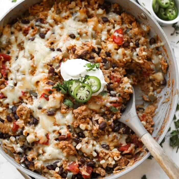 Skillet filled with cheesy rice, black beans, corn, tomatoes, and ground meat, topped with sour cream and sliced jalapeños, served with a wooden spoon.