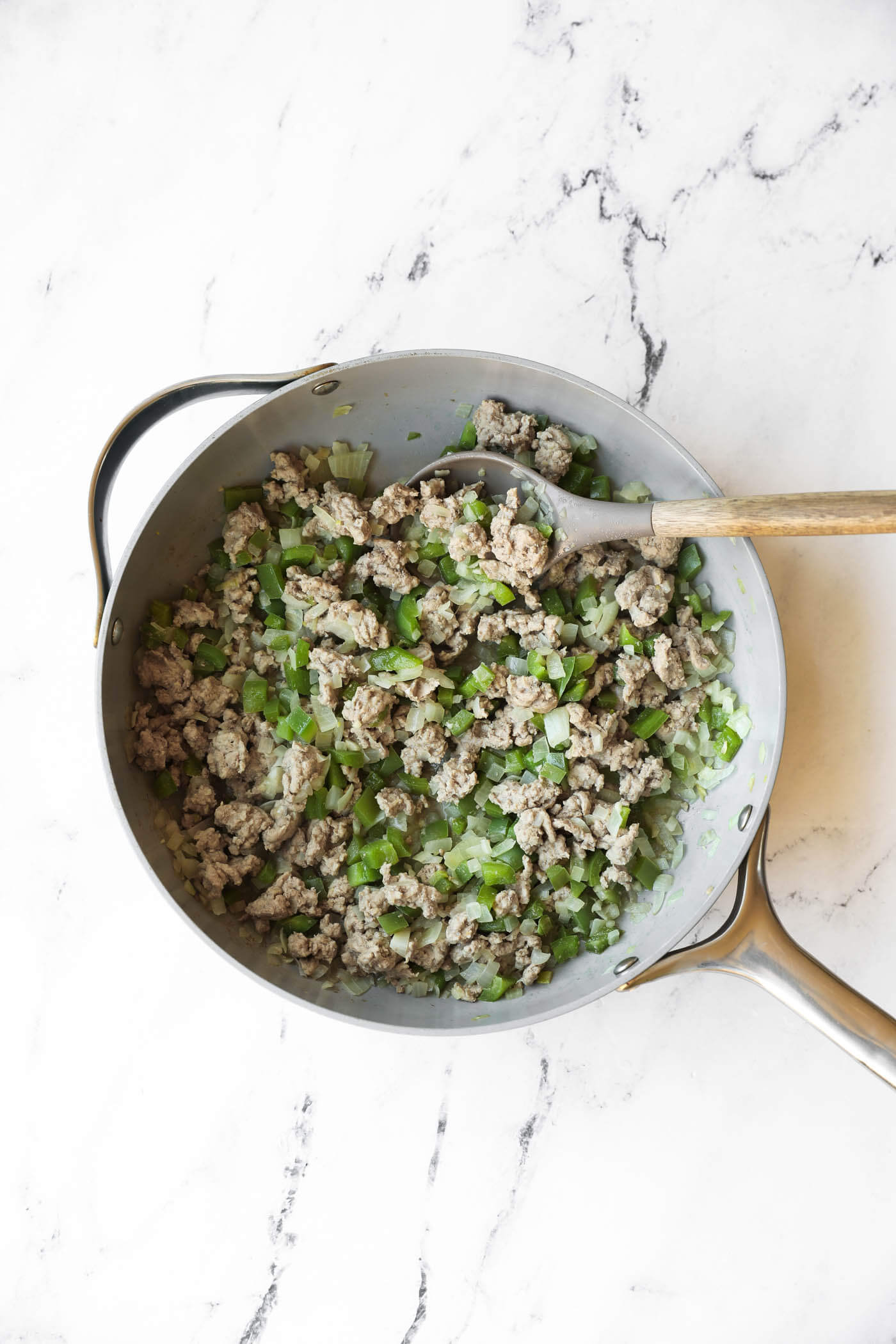 A metal skillet with cooked ground meat, chopped green bell peppers, and onions, with a wooden spoon resting inside on a white marble surface.