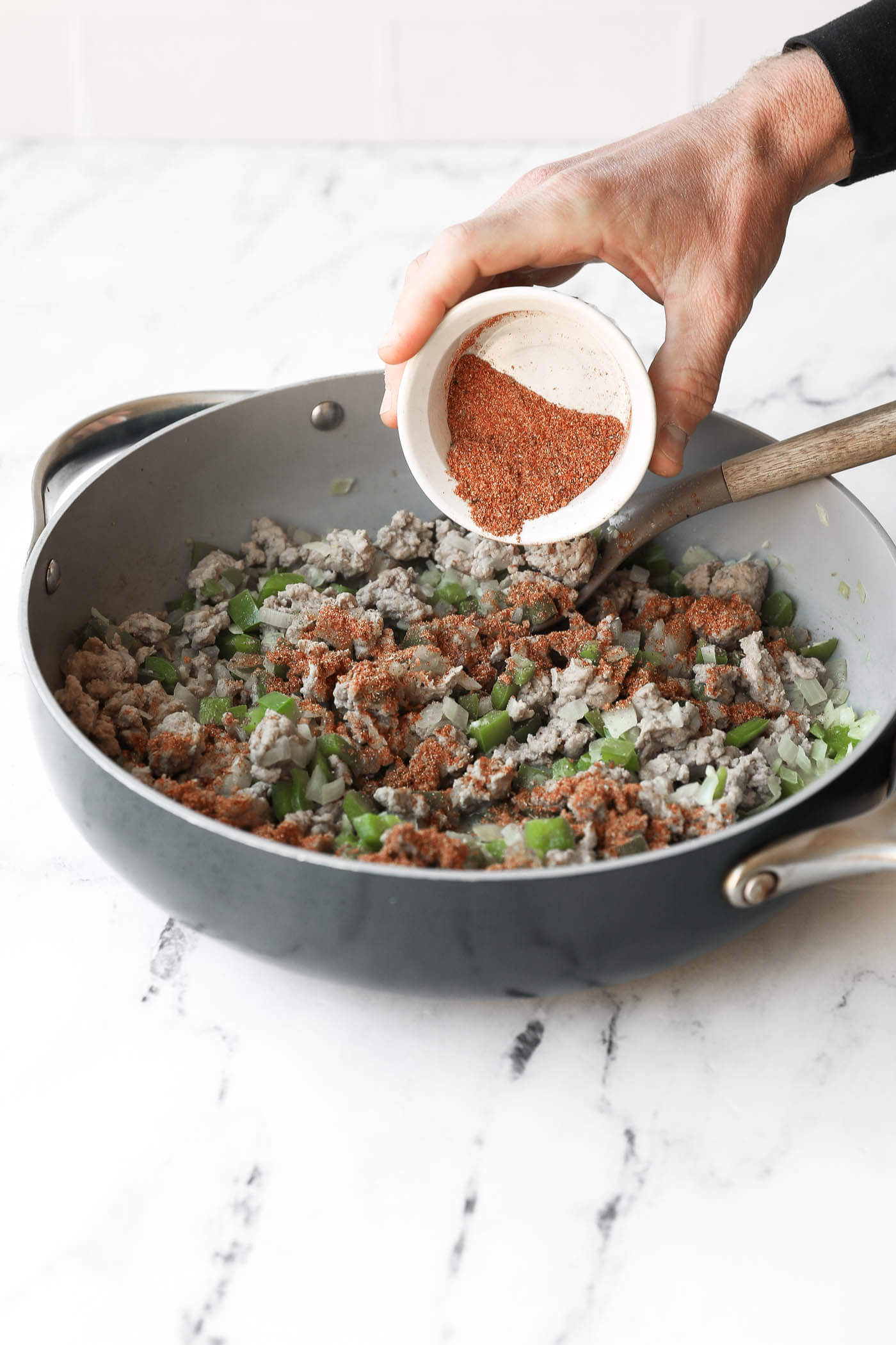 A hand pours taco seasoning from a small bowl into a skillet with cooked ground meat, onions, and green bell peppers on a marble countertop.
