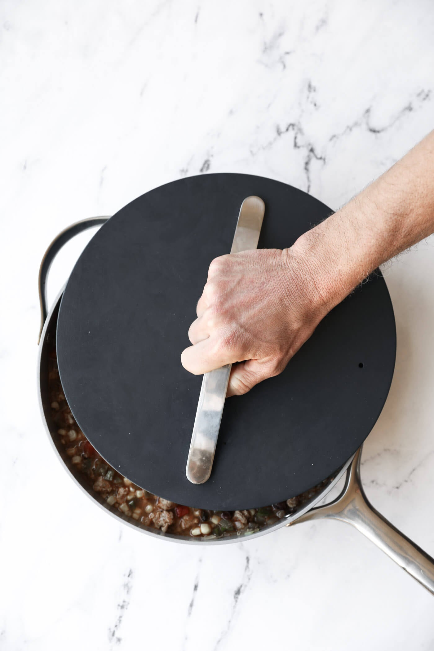 A hand holding a metal handle on a black lid covering a saucepan with food inside, placed on a white marble surface.