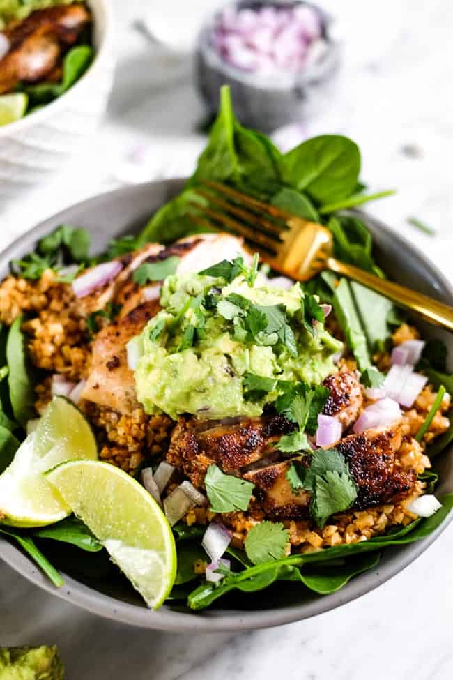 Angled shot of chicken burrito bowl, topped with chopped red onion, guacamole, cilantro and lime wedges. 