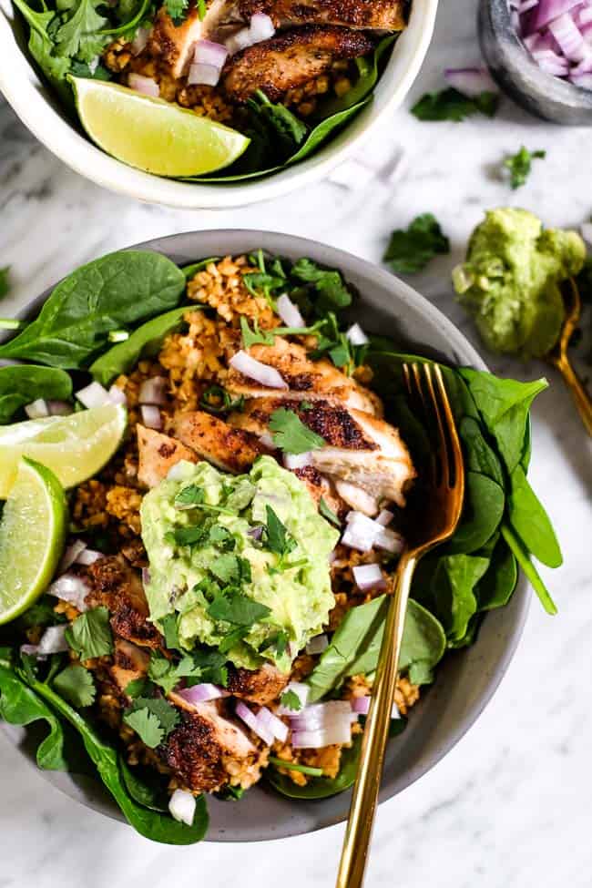 Chicken burrito bowl served up with chopped red onion, guacamole, cilantro and lime wedges in a bowl with fork. 