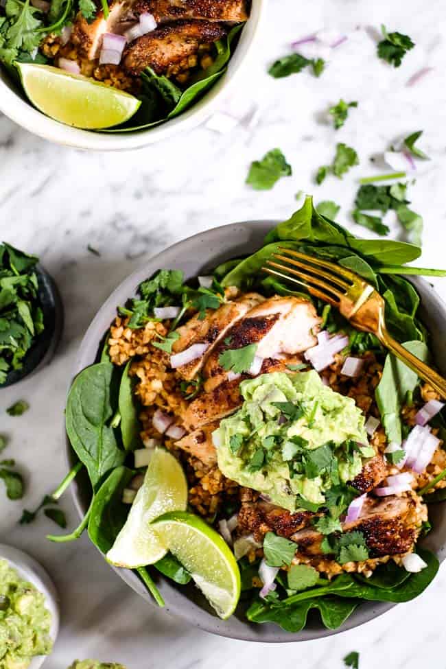 Chicken burrito bowl with chopped red onion, guacamole, cilantro and lime wedges with a fork. 