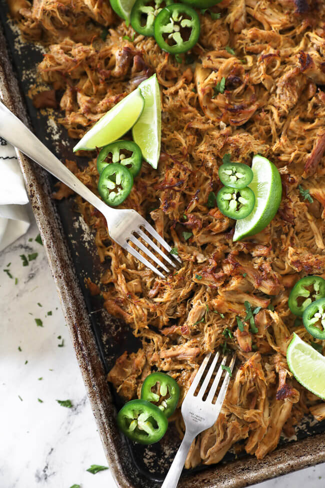 Overhead vertical image of crispy chicken carnitas on a sheet pan with two forks for shredding, lime wedges, sliced jalapeño and chopped cilantro. 