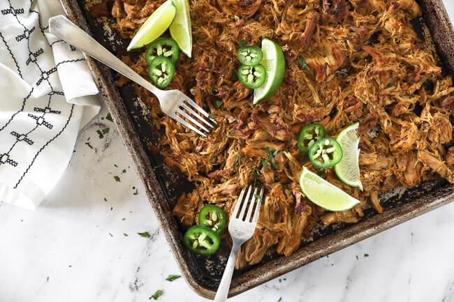 Overhead horizontal image of crispy, shredded chicken carnitas on a sheet pan with two forks, lime wedges, jalapeño slices and fresh chopped cilantro.  