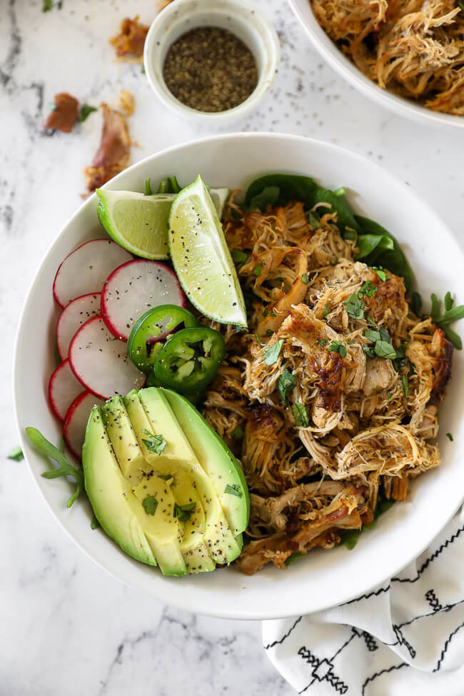 Overhead vertical image of chicken carnitas served in a bowl with greens, sliced radish, lemon wedges, sliced jalapeño and avocado slices. Topped with chopped cilantro. 
