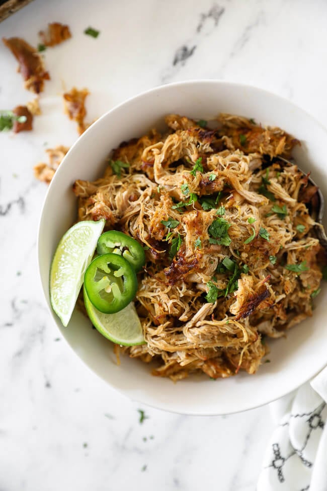 Overhead vertical image of chicken carnitas in a bowl with garnish of lime wedges, sliced jalapeño and chopped cilantro. 