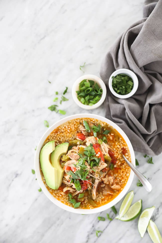 Zoomed out image of chicken fajita soup in a bowl with a spoon in it. Topped with chopped green onion, cilantro and sliced avocado. Lime wedges and extra green onion and cilantro on the side. 