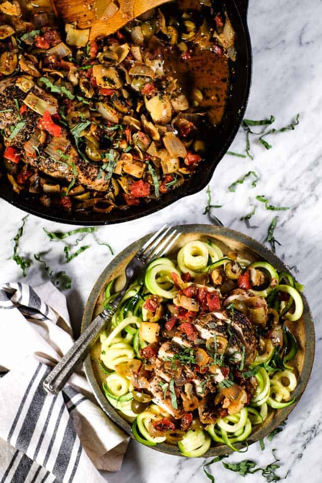 Chicken in tomato sauce served on a plate over zoodles with fresh basil on top. Skillet of chicken and sauce in background. 