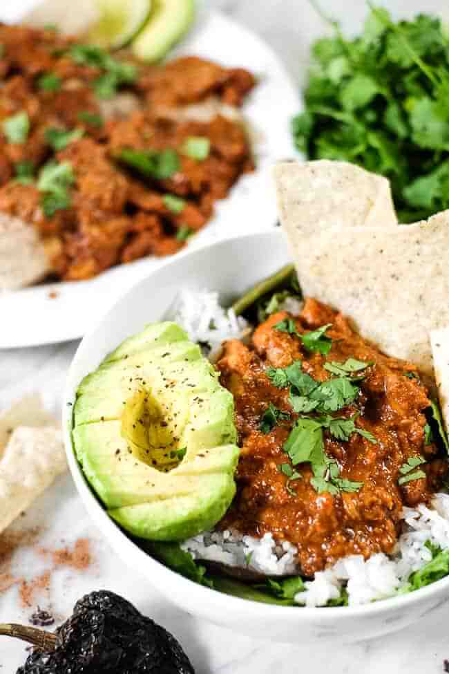 Chicken mole recipe in a bowl with greens, rice, avocado, grain free tortilla chips and chopped cilantro. Enchiladas in the background. 
