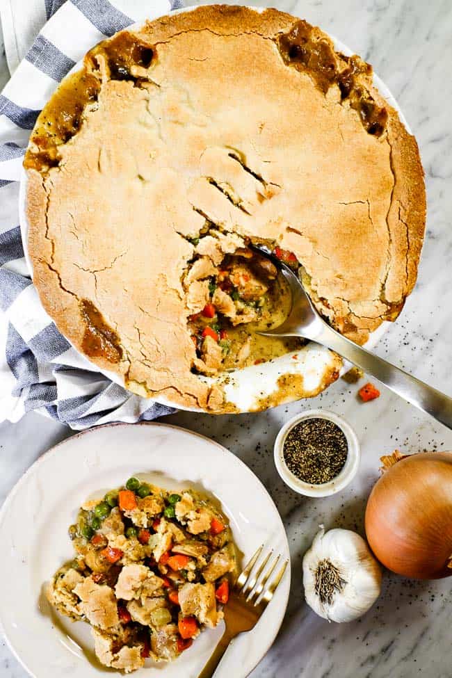 Chicken pot pie casserole served up on a plate with a fork. Casserole in background with a blue striped towel and serving spoon in dish. Further out shot of whole casserole. 