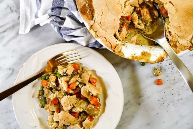 Chicken pot pie casserole served up on a plate with a fork. Casserole in background with a blue striped towel and serving spoon in dish. 