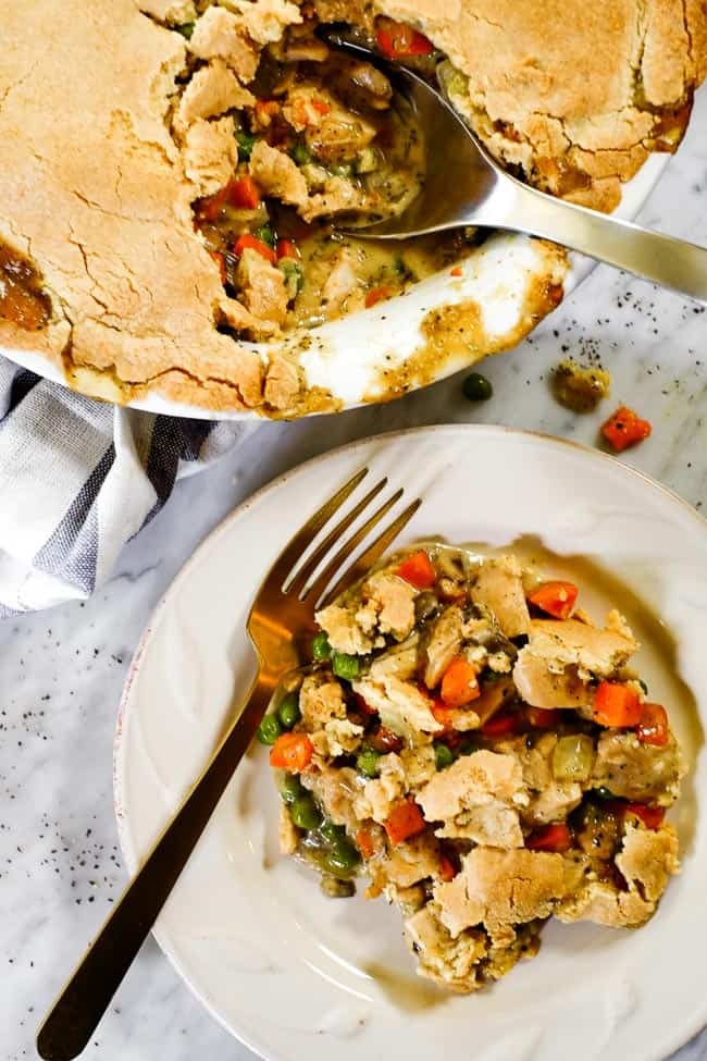 Chicken pot pie casserole served up on a plate with a fork. Casserole in background with a blue striped towel in the background with a serving spoon in dish. 