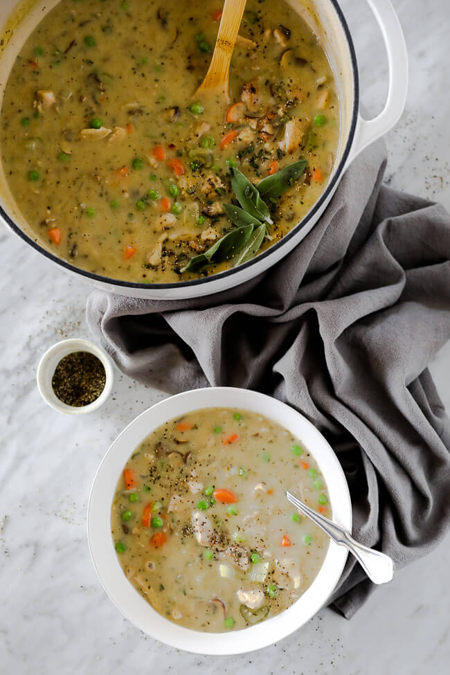 Vertical image of chicken pot pie soup served in a bowl with a spoon in it. Large soup pot in the image as well with ground pepper on the side. 