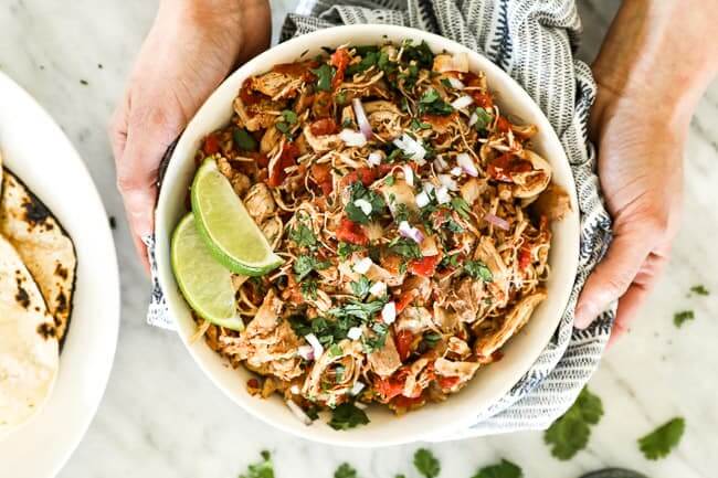 Holding a bowl of shredded mexican chicken topped with chopped cilantro, red onion and lime wedges. 