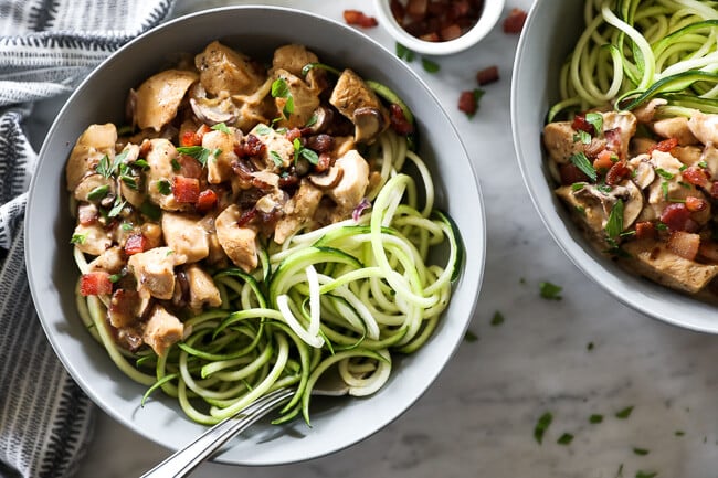 Creamy mushroom chicken served over zoodles in a bowl with a fork dug in. Extra bacon bits on the side. 