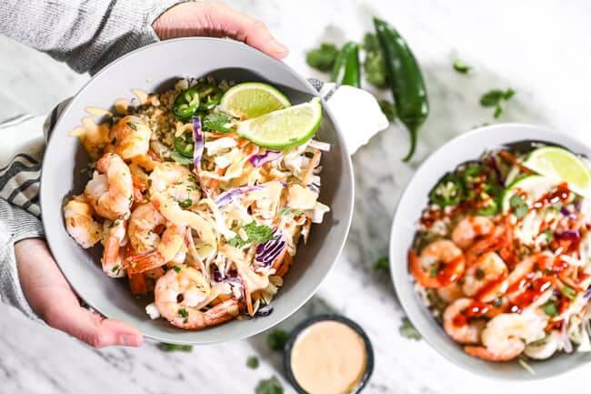 Holding a cilantro lime shrimp bowl over table with extra bowl, sauce and jalapeño in background. Bowls are topped with sauce, jalapeño slices and lime wedges. 