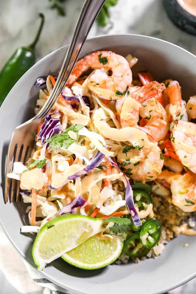 Close up shot of cilantro lime shrimp bowl with fork in it, topped with sauce, jalapeño slices and lime wedges. 