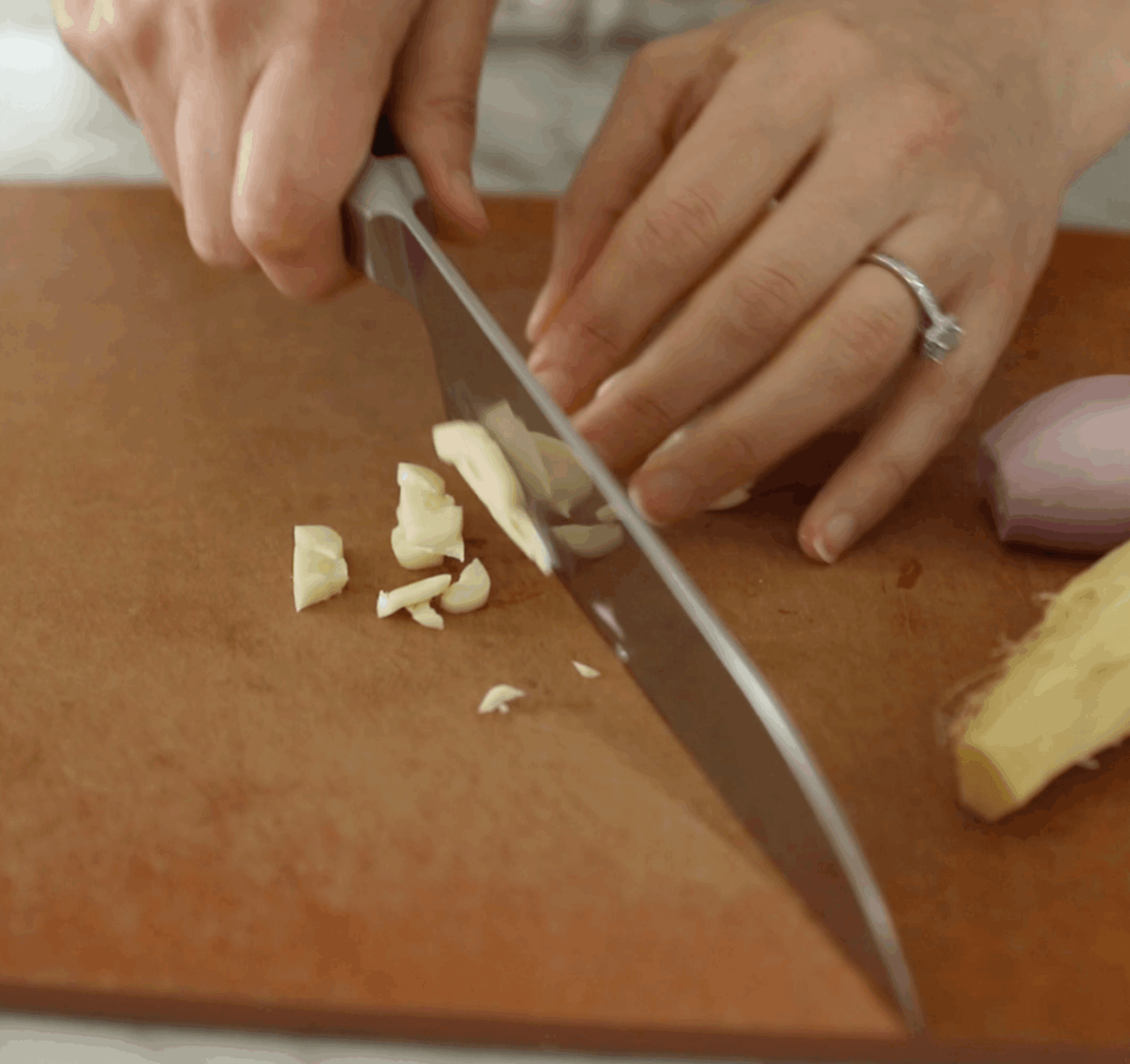 A person chops garlic on a brown cutting board with a large kitchen knife; a shallot and ginger are also on the board.
