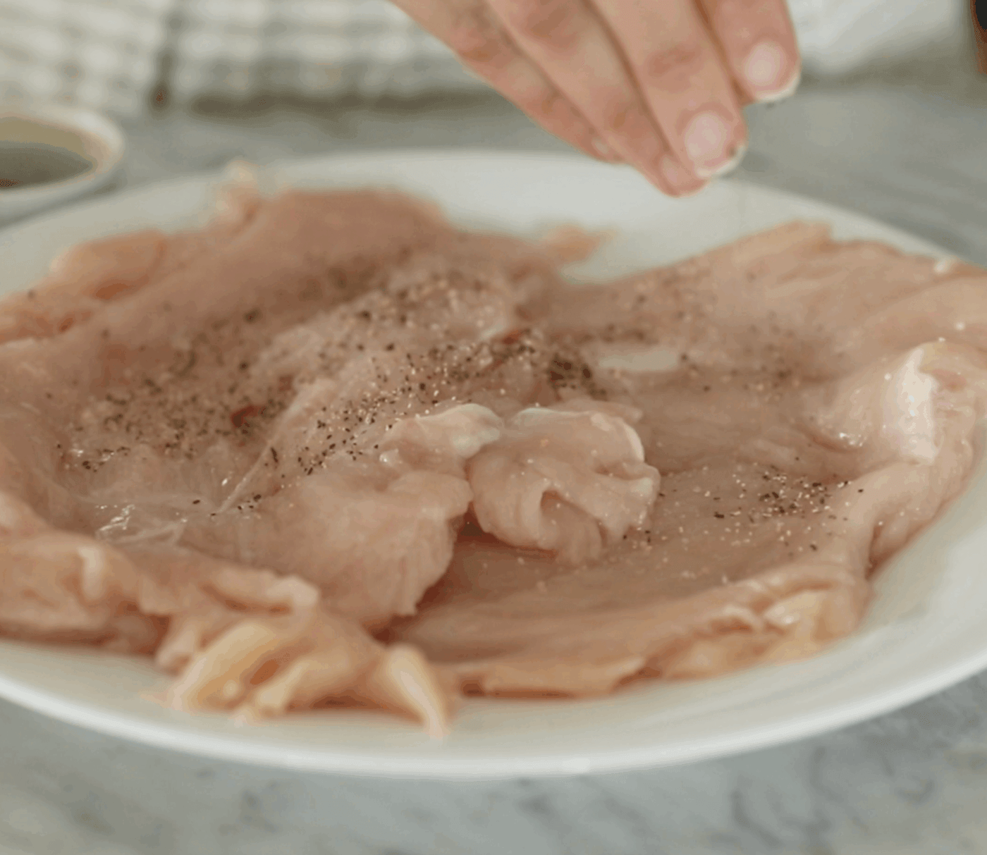 A hand sprinkles seasoning onto raw chicken breasts on a white plate placed on a marble countertop.