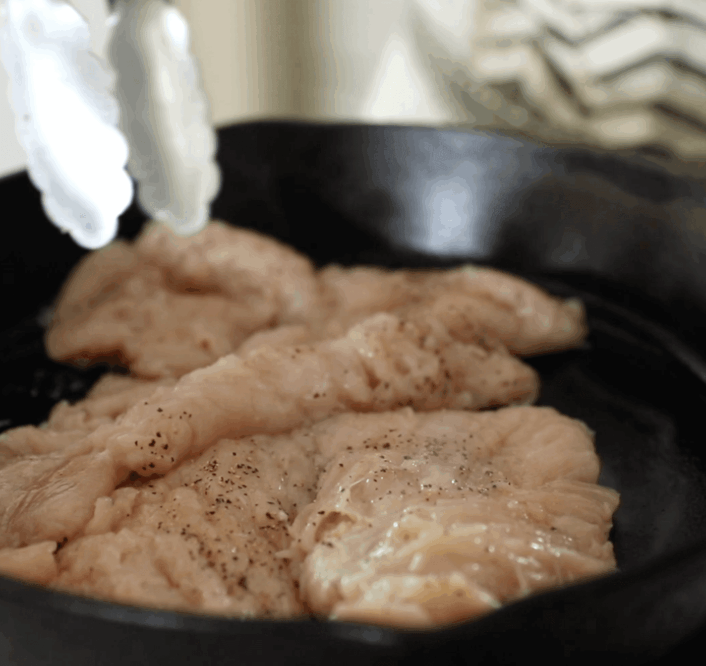 Raw, seasoned chicken breasts being placed into a black cast iron skillet with metal tongs.