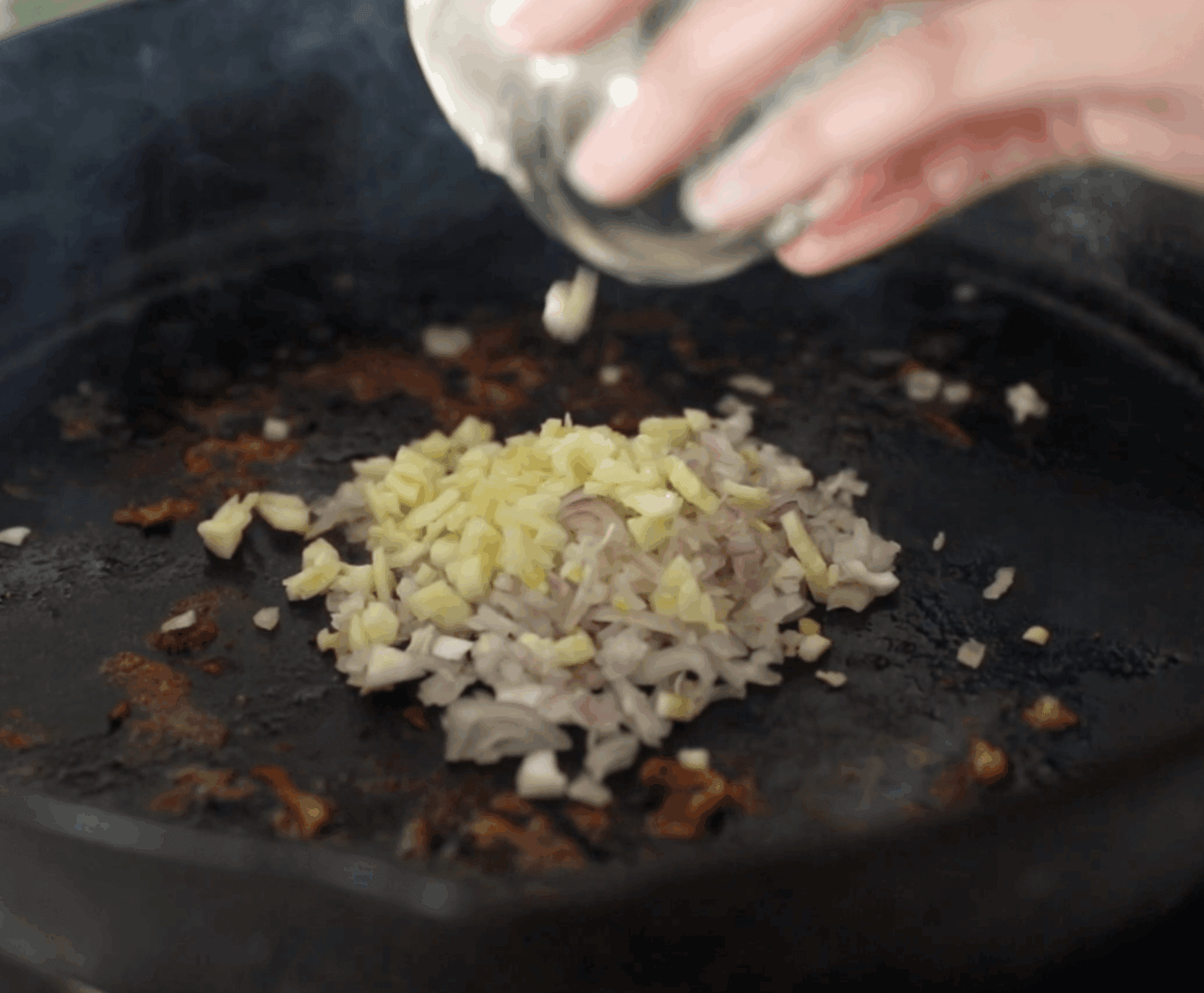Chopped onions and garlic are added from a small bowl into a black cast iron skillet for cooking.