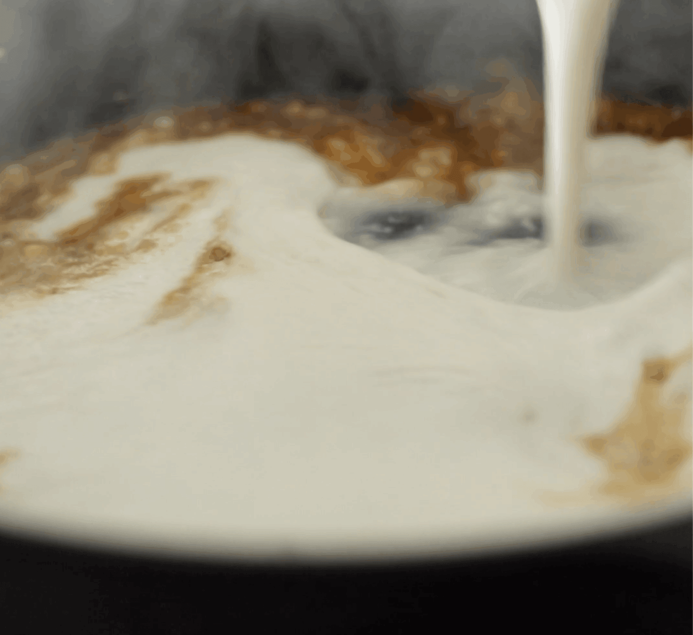 A close-up of milk being poured into a pot of simmering liquid, causing the liquid to swirl and mix.