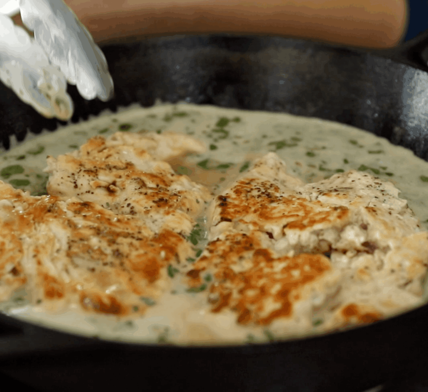 Chicken breasts cooking in a creamy sauce with herbs in a black skillet, with metal tongs visible above the pan.