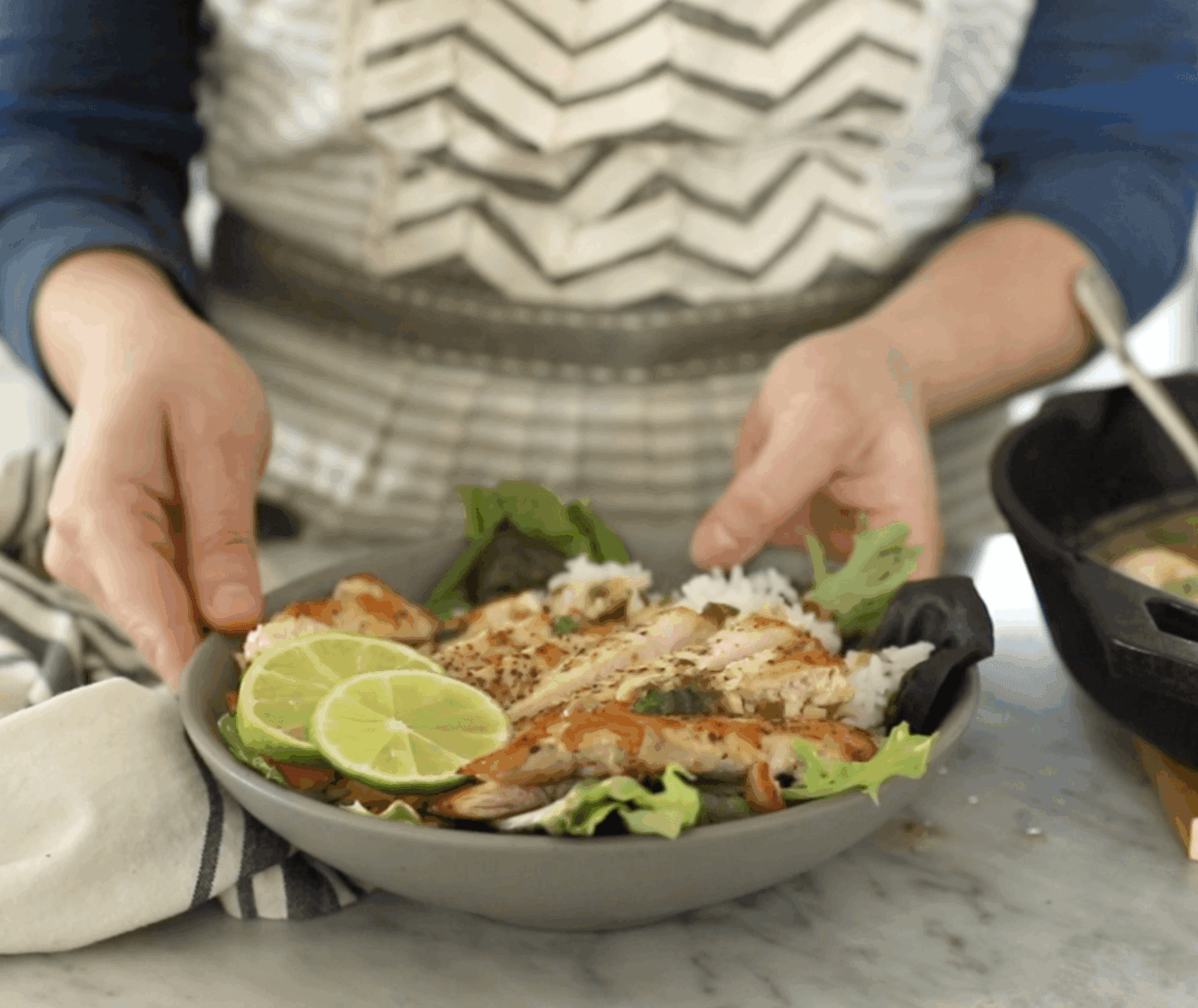 A person in a striped apron holds a plate of coconut lime chicken on rice, garnished with lettuce and lime slices, next to a pot on a marble countertop.