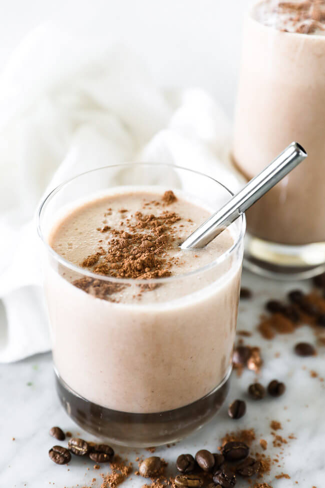 Vertical angled image of coffee smoothie in a glass with cacao sprinkled on top and a stainless steel straw. 