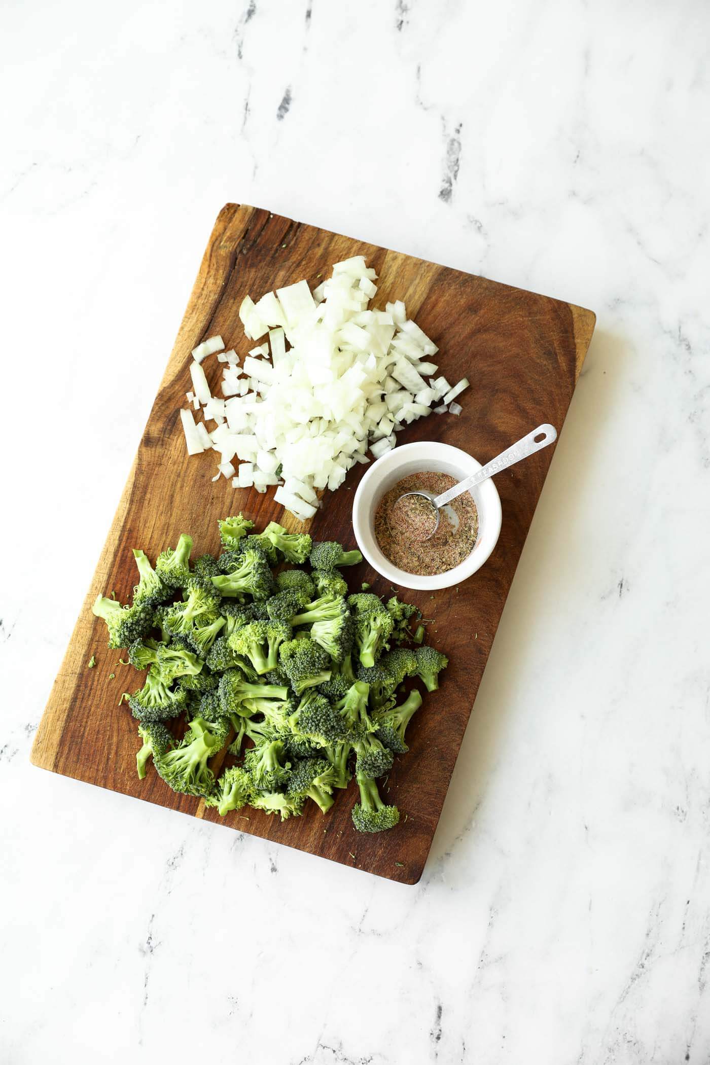 Chopped onion, broccoli florets, and a small bowl of mixed seasoning on a wooden cutting board on a marble surface.