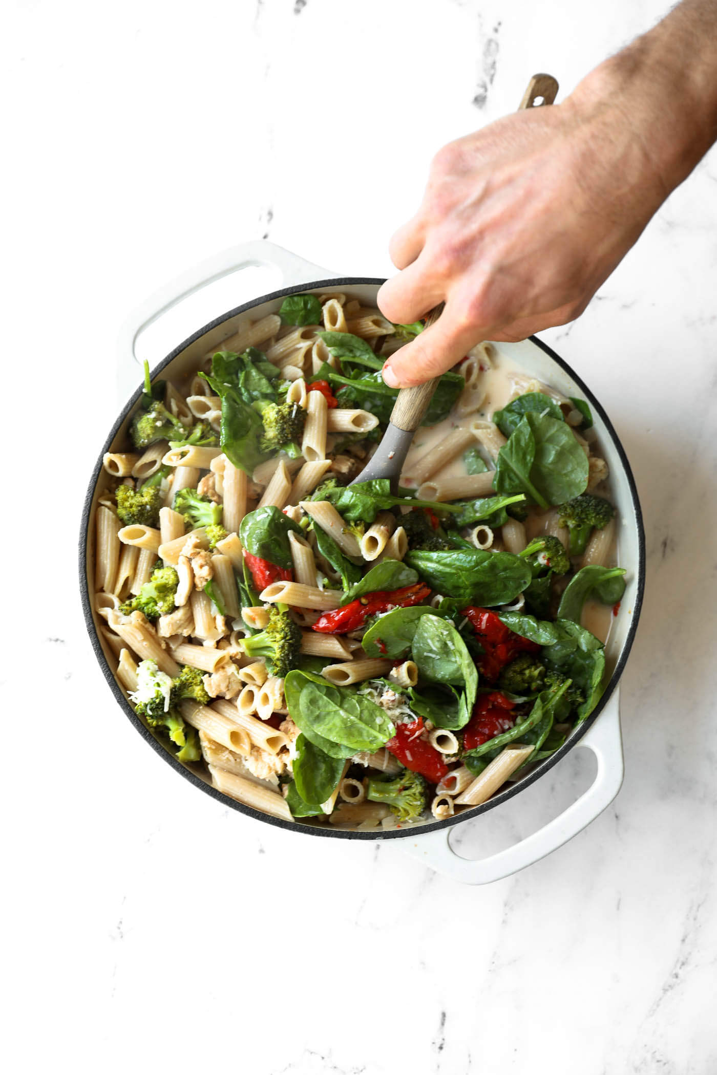 A hand stirs penne pasta, spinach, broccoli, and sun-dried tomatoes in a white pot with a creamy sauce on a white surface.