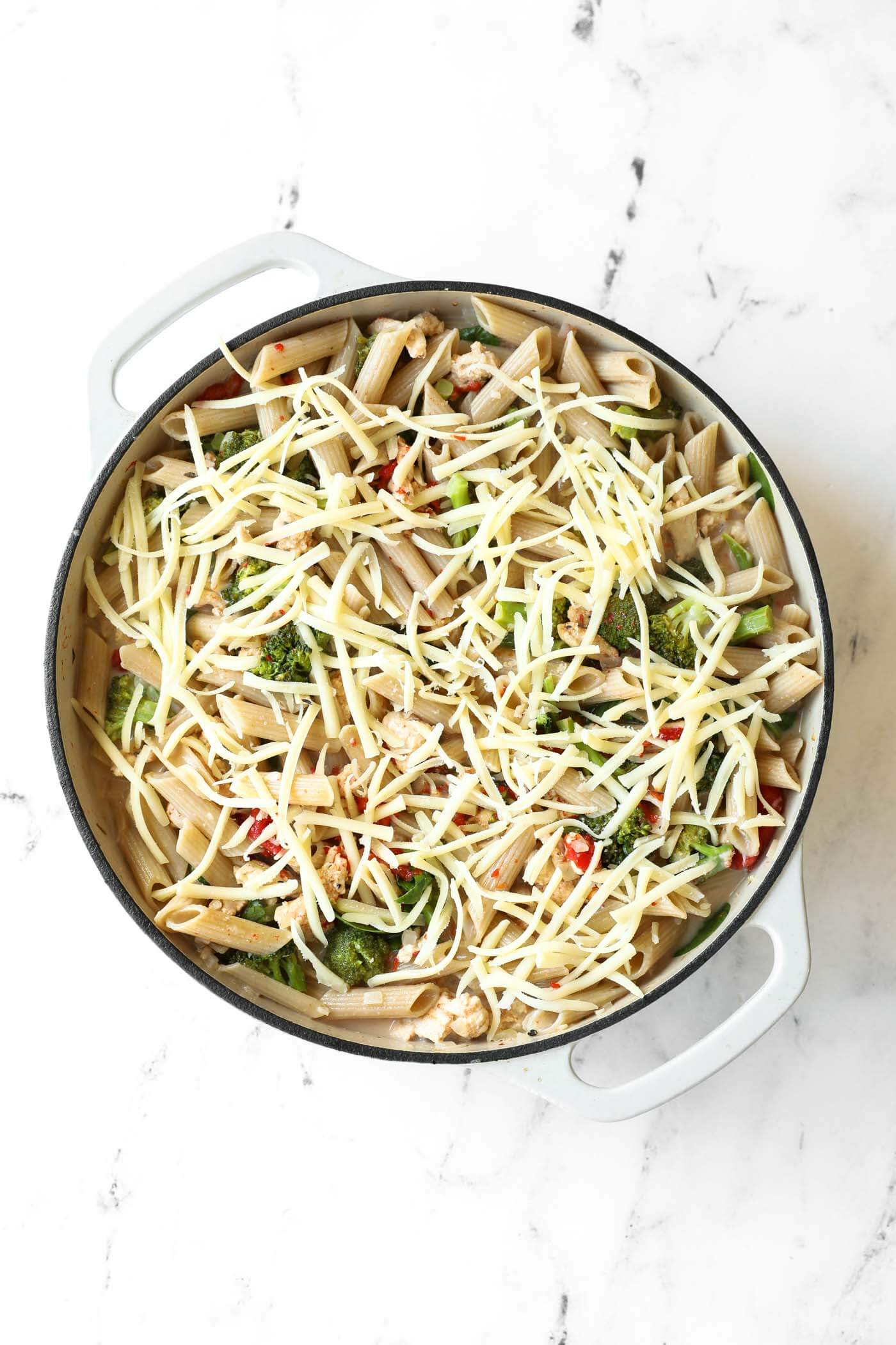 A white pot filled with penne pasta, vegetables, and cooked ground meat, topped with shredded cheese on a marble surface.
