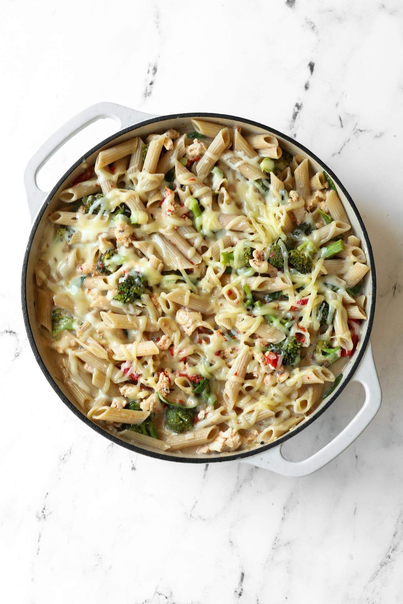 A round white casserole dish filled with penne pasta, chopped vegetables, and melted cheese, set on a white marble surface.