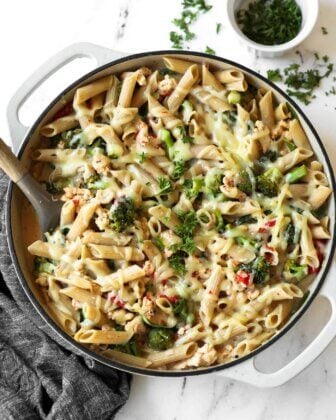 A pan of baked penne pasta with melted cheese, broccoli, and herbs, with a spoon and a small bowl of chopped parsley nearby.
