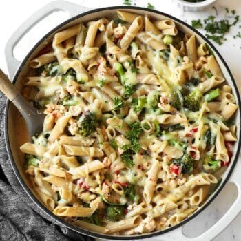 A pan of baked penne pasta with melted cheese, broccoli, and herbs, with a spoon and a small bowl of chopped parsley nearby.