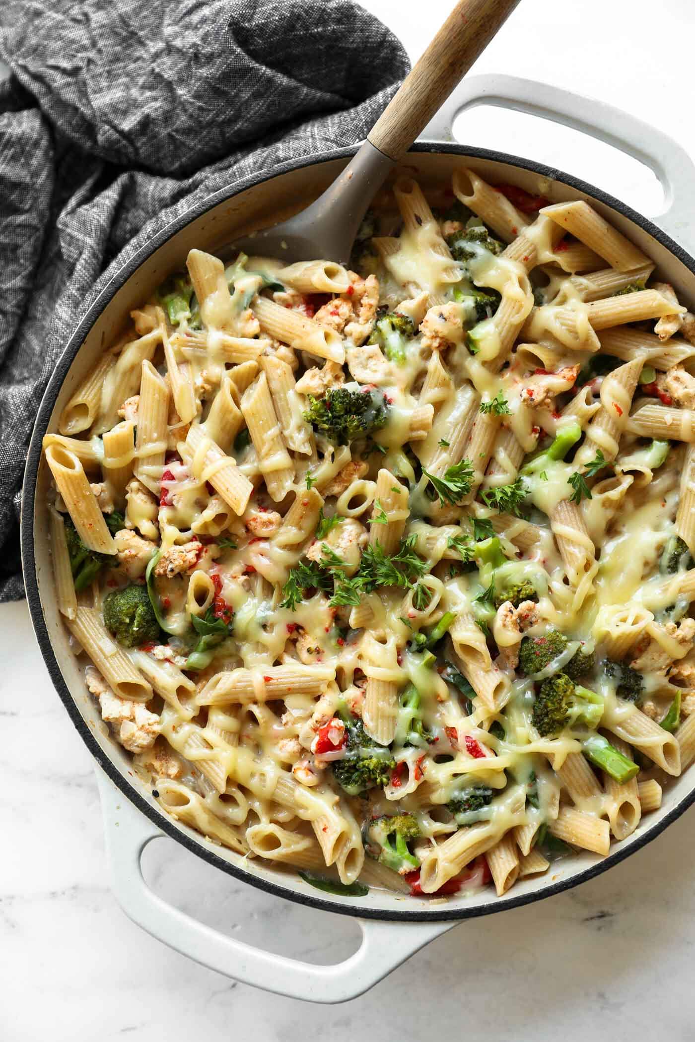 A pan of baked penne pasta with melted cheese, broccoli, and herbs, with a wooden spoon resting inside.