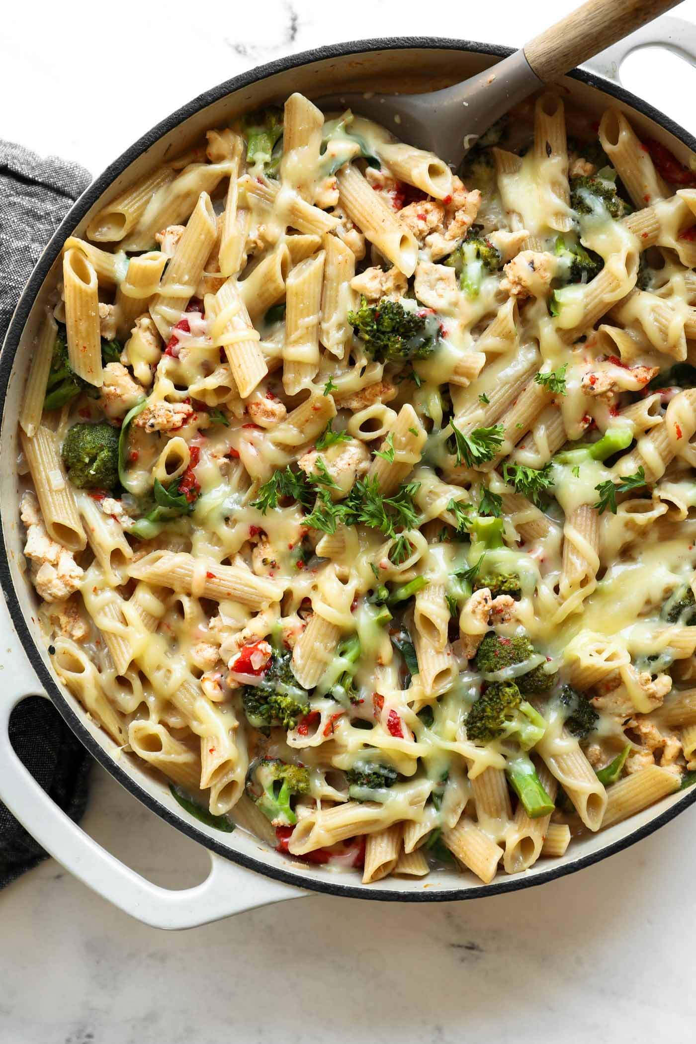 A skillet filled with baked penne pasta, broccoli, melted cheese, and herbs, with a wooden-handled serving spoon resting inside.
